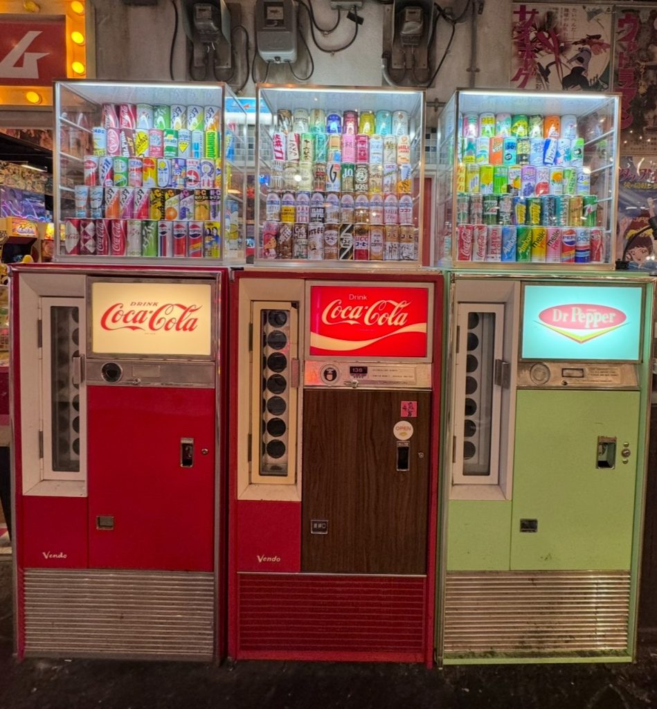 Three retro vending machines with a wide variety of drinks offered. Two of the machines are labeled as “Coca-Cola” and the other as “Dr. Pepper.”