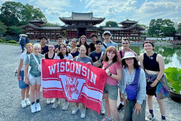 Smiling students pose in front of a Japanese temple while holding a red and white Wisconsin flag.