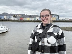 Caucasian woman with brown hair pulled into a pony tail. She is wearing glasses and a black and white plaid jacket. In the background is a row of colorful houses along a waterfront.