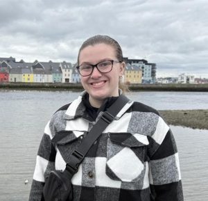 Caucasian woman with brown hair pulled into a pony tail. She is wearing glasses and a black and white plaid jacket. In the background is a row of colorful houses along a waterfront.