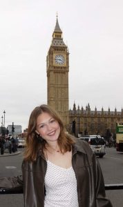 Caucasian woman with medium length brown hair wearing a black pleather jacked and a white shirt. She is standing in front of the Big Ben clock tower in London, England.