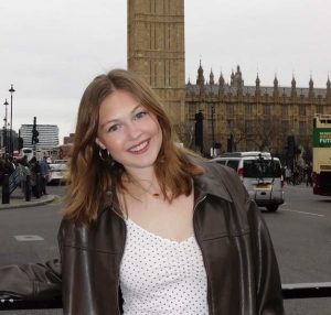 Caucasian woman with medium length brown hair wearing a black pleather jacked and a white shirt. She is standing in front of the Big Ben clock tower in London, England.