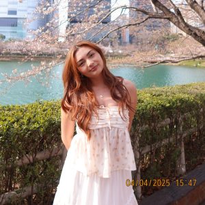 A young woman with long brown hair wearing a white flowy top and skirt stands in front of a lake lined with cherry blossom trees. Behind her, tall modern buildings rise under a clear blue sky. The timestamp “04/04/2025 15:47” is visible at the bottom.