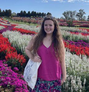 A woman with long wavy hair wearing a pink sleeveless top and a floral skirt poses in front of a vast field of colorful flowers arranged in striped rows of red, white, and purple. The sky above is clear and bright blue with scattered clouds.
