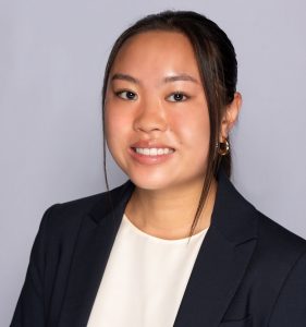 A young woman with straight dark hair tied back, wearing a navy blazer over a white top, smiling in front of a plain light-gray background.