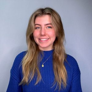 A young woman with long blonde hair, wearing a bright blue sweater and a gold necklace, smiling in front of a plain gray background.