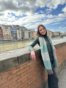 A smiling woman wearing a green top, black pants, and a patterned scarf leans casually against a brick wall along a riverfront. Colorful buildings line the opposite bank under a partly cloudy blue sky.
