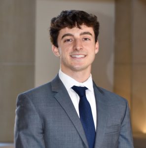 A young man with short dark wavy hair, wearing a gray suit with a white shirt and navy tie, smiling in front of a blurred indoor background.