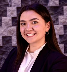 A young woman with straight dark brown hair, wearing a black blazer over a white blouse, smiling in front of a textured gray stone wall background