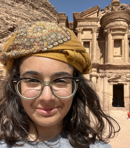 A woman wearing glasses and a mustard-yellow embroidered headscarf smiles at the camera. She is outdoors in bright sunlight, standing in front of the ancient carved stone façade of Petra in Jordan.