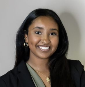 A young woman with long dark hair, wearing a black blazer over a green blouse and gold jewelry, smiling in front of a plain light-colored background.