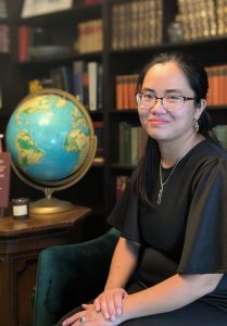 A woman with glasses and long dark hair, dressed in a black outfit, sits on a green chair in front of bookshelves filled with books. A globe on a wooden side table is prominently visible beside her.