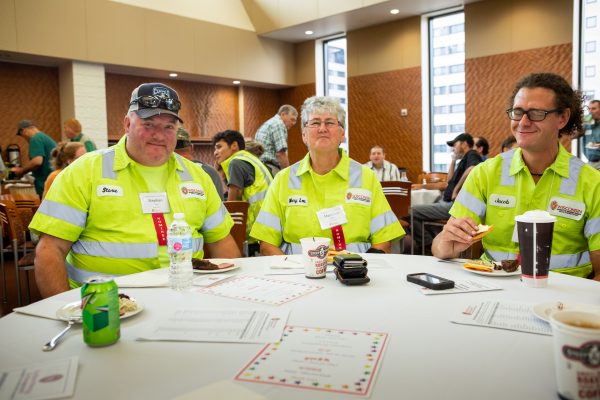 three people in bright yellow safety uniforms sit around a round table with white tablecloth with various expressions on their faces