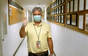 Hector is standing in long hallway with awards and photos in frames on the wall. He is giving a thumbs up and wearing a face covering