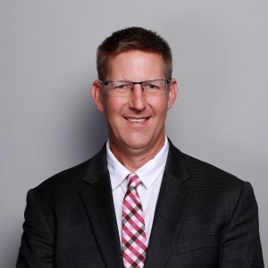 person standing against a rough tan brick wall wearing glasses, a collared shirt with pullover and smiling