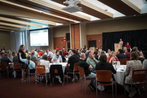 people seated in a banquet hall talking to each other at tables while a slideshow is played on large screens in the background