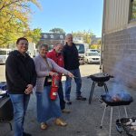 four people stand outdoors near a building with grills smoking and cooking food