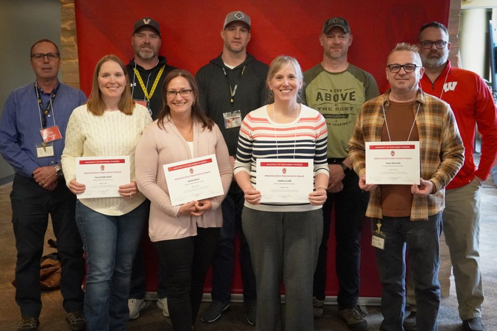 a group of 9 employees pose with award certificates smiling against a red and stone background in a hallway