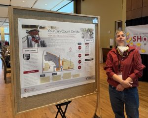 A person in a red shirt and jeans, with a mask and a name badge around their neck, stands next to a bulletin board displaying a poster that says "You Can Count On Me," and has an image of a bicycle.
