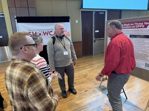 A man in red stands in front of a poster, speaking with three people -- a man in a plaid shirt, a woman in stripes, and another man in a gray sweater, smiling.