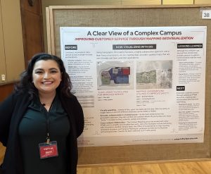 woman wearing a black shirt with a name badge stands next to a bulletin board, where a poster is pinned. The poster says "A Clear View of a Complex Campus" with maps.