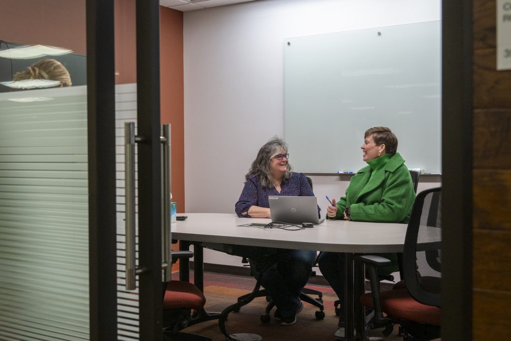 Two people viewed from outside a conference room door as they sit at an oval table side-by-side looking at one another and talking while smiling in front of a laptop