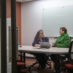 Two people viewed from outside a conference room door as they sit at an oval table side-by-side looking at one another and talking while smiling in front of a laptop