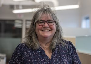 Person with shoulder-length gray hair, dark-rimmed glasses and a navy blue shirt with pink and white polka dots smiles widely standing in an office setting