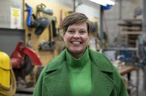 Person with light brown short hair parted at the side wearing gold-colored hoop earrings and a vibrant green turtleneck and green coat smiles widely while standing in front of a workshop with metal parts, yellow hose, and red machinery