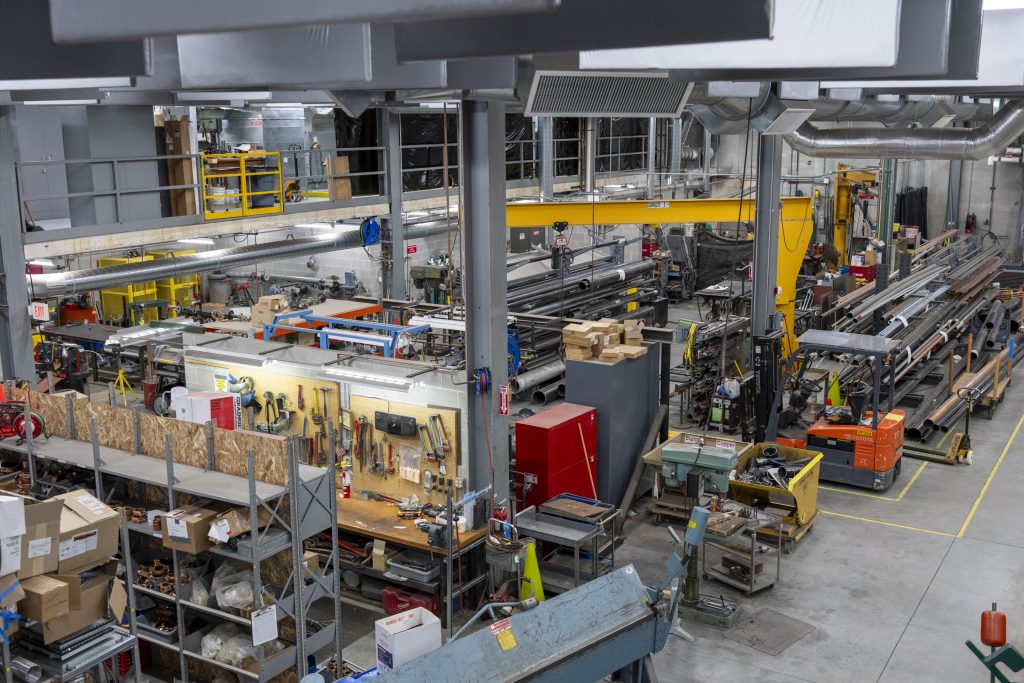 Wide view from above a two-story indoor machinery shop space constructed with large gray metal beams attaching the bottom floor to the roof of the space. Bright yellow metal machinery, floor tape, storage and gates stand out. Shop supplies are seen, storage racks, cabinets and many tools