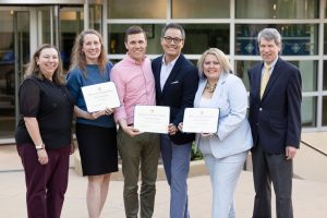 6 recipients standing together holding three pieces of paper that are rewards
