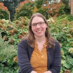 Photo of a woman wearing glasses, a goldenrod shirt and gray cardigan, sitting in front of foliage changing from green to autumn colors.