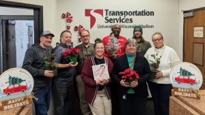 *Eight people pose for photo, two happy holidays signs on surrounding surfaces, participants hold red flowers, a small tree and a picture with a candy cane*
