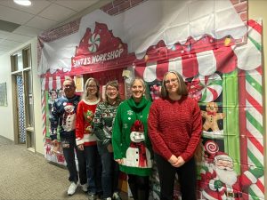 *Five people pose for photo in holiday sweaters, standing in front of "Santa's Workshop" themed sheet on wall, colorful and festive*