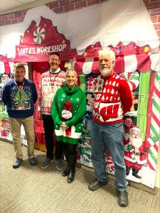 *Four people in holiday sweaters pose in front of "Santa's Workshop" sheet on wall*