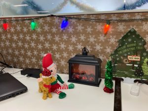 *Close up, wrapping paper with snowflakes on short wall, colorful string lights, elf and reindeer toy on desk, small tinsel tree with star, small plastic fireplace, tree cutout* 