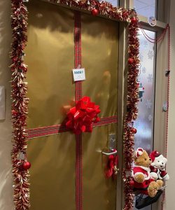 *Door wrapped like a present in gold wrapping paper and a red ribbon, tinsel on doorframe, two stuffed animals sit in window*