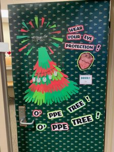*Door wrapped in green wrapping paper with trees on it, large red and green paper tree with safety goggles at top, reads "wear your eye protection, O' PPE tree! O' PPE Tree!"*
