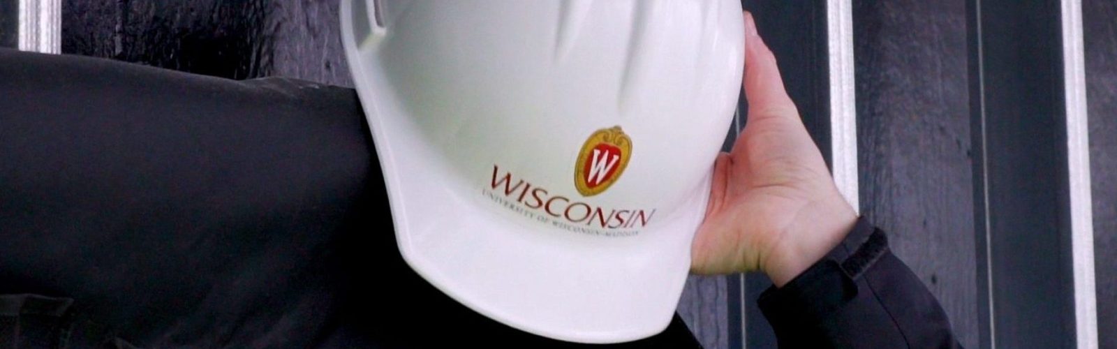 A hard hat representing construction workers at UW-Madison is being held up against a dark backdrop.