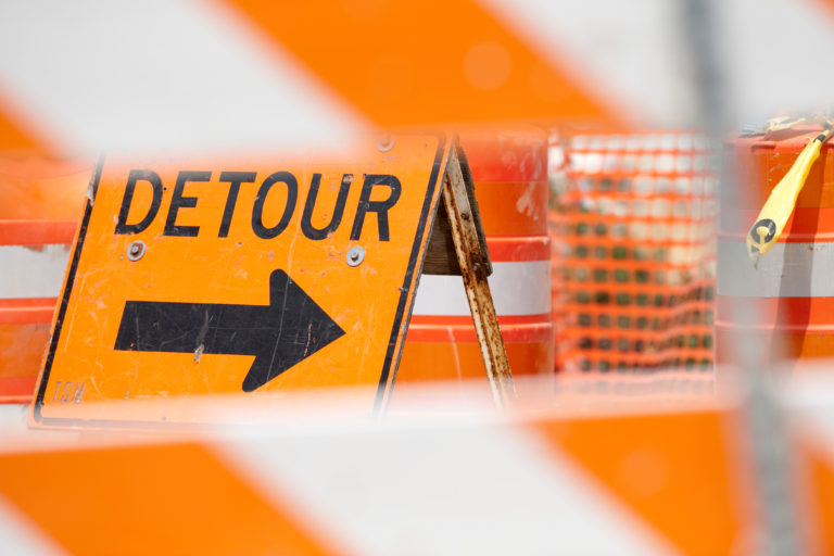 Picture of an area blocked by construction barricades and other traffic management signs. Focus is on a bright orange sign stating "Detour" with an arrow pointing to the right.