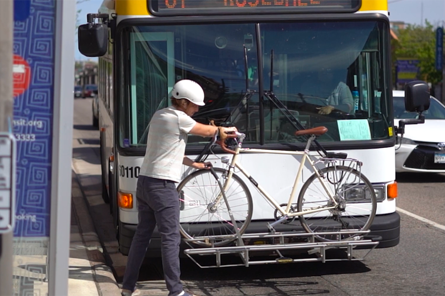 Photo of male-presenting individual wearing a white bicycle helmet loading a bicycle onto a front-mounted bicycle rack on a city bus.