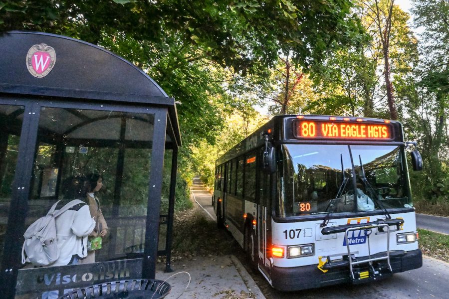 Photo of a city bus stopped at a covered bus shelter in a wooded area where two people are waiting to board.