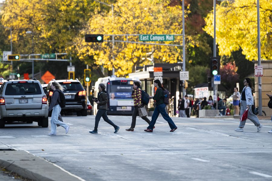 Photo of pedestrians on a crosswalk on a busy street with yellow fall foliage in the background.