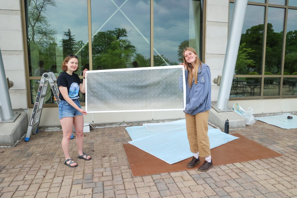 Two individuals holding up a large, clear panel with small white dots on it. They are standing outdoors in front of a glass building, with a ladder and other supplies nearby.