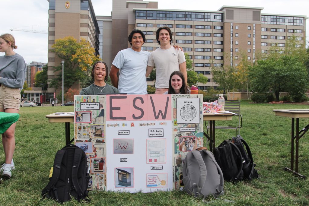 Four students standing and sitting behind a table with a display board that reads "ESW." They are outdoors on a grassy area with buildings in the background.