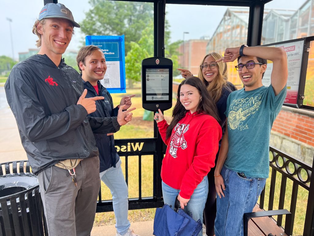 A group of five smiling people standing at a bus stop, pointing at a digital display screen. The group appears excited, and the setting is outdoors with a building visible in the background.