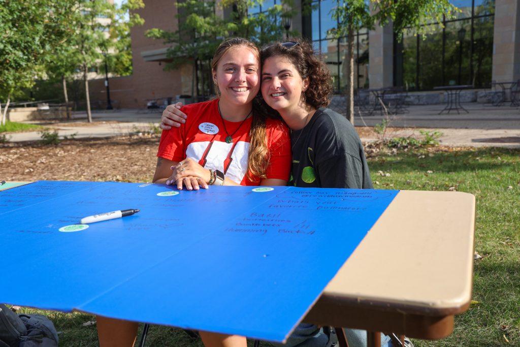 Two individuals seated at a table outdoors, smiling and leaning toward each other. A large blue poster with handwritten text is on the table.