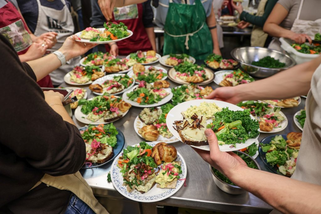 A close-up of several plates of colorful, fresh food being served in a kitchen setting. Hands are visible holding plates and serving food.