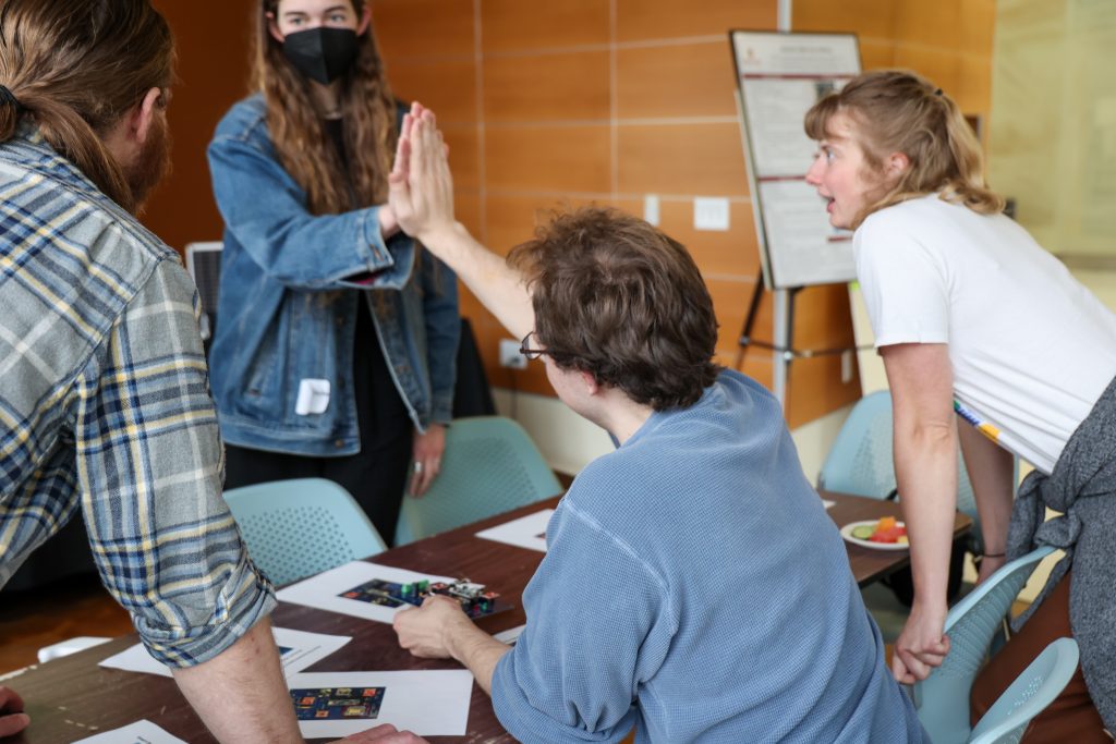 Four people gathered around a table indoors, engaged in a collaborative activity. One person is high-fiving another, while others look on with interest.