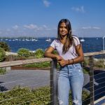 Maisha Islam poses, dressed casually in front of of a lake with boats and a blue sky